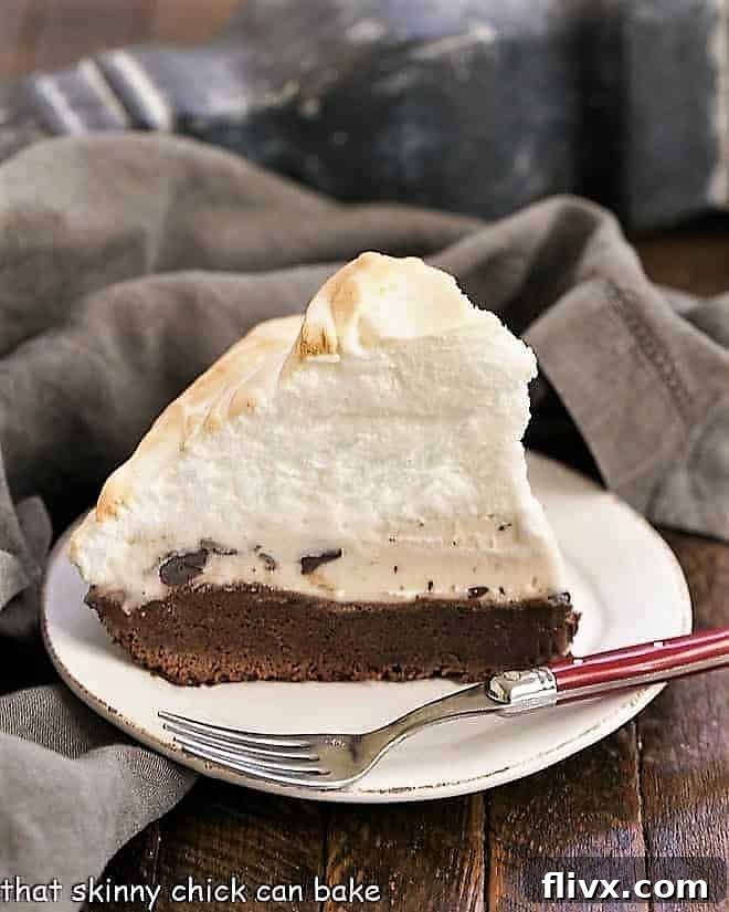 A perfectly sliced piece of Brownie Baked Alaska resting on a white dessert plate, showing the distinct layers of brownie, melting ice cream, and crisp, toasted meringue.