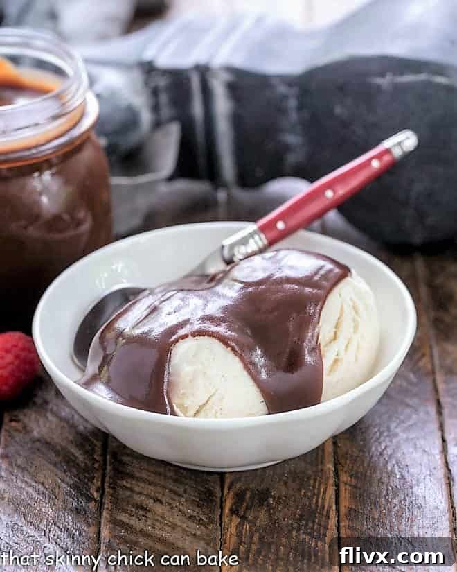 Fudgy Chocolate Sauce you can make at home! The ultimate topping for a perfect bowl of ice cream. A charming white bowl filled with creamy vanilla ice cream, generously topped with glistening, dark hot fudge sauce, with a red-handled spoon resting beside it.