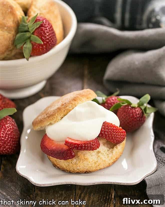 Three perfectly assembled Strawberry Shortcakes with White Chocolate Mousse on a square white plate, with a bowl of unbaked biscuits visible in the soft background.
