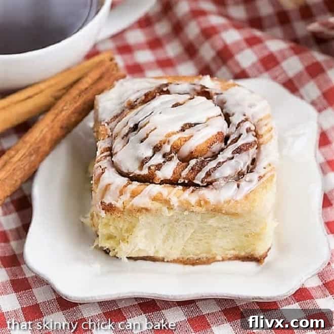 The Best Cinnamon Buns on a square white plate with cinnamon sticks on a red and white checked napkin, highlighting their perfect golden-brown exterior and generous glaze.