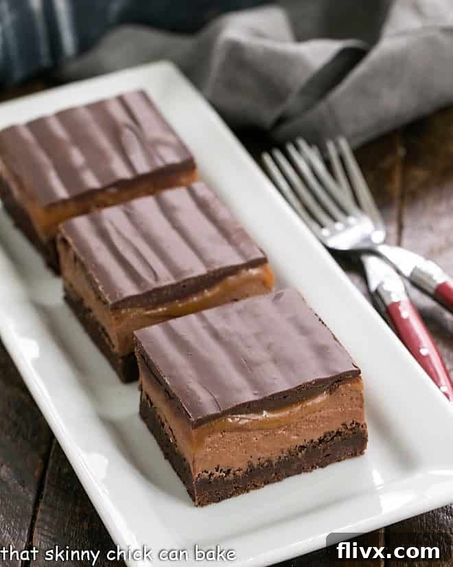 Three decadent Milky Way Brownies artfully arranged on a pristine white ceramic tray, showcasing their luxurious layers of chocolate, nougat, and caramel.