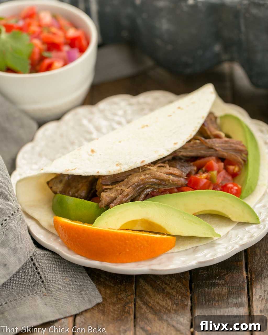 Delectable slow cooked Pork Carnitas nestled in a soft tortilla, elegantly presented on a white plate with fresh avocado slices and orange garnishes.