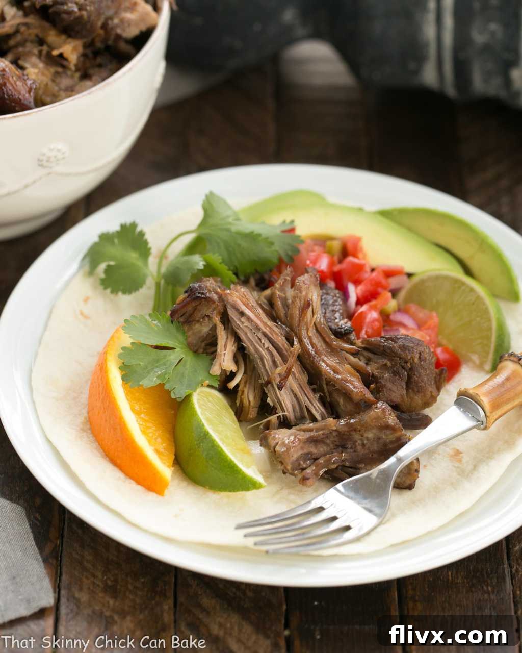 A vibrant display of slow cooked Pork Carnitas presented on a white plate, artfully garnished with fresh tomatoes, creamy avocado, and bright citrus slices, highlighting its fresh appeal.