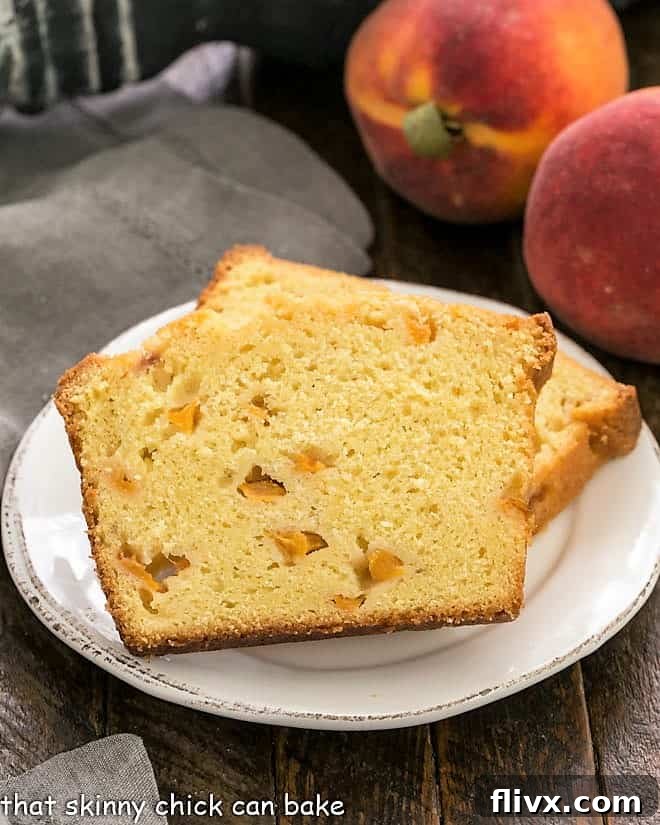 Two slices of peach cake on a white dessert plate with 2 peaches in the background, showcasing the moist crumb and juicy peach pieces.
