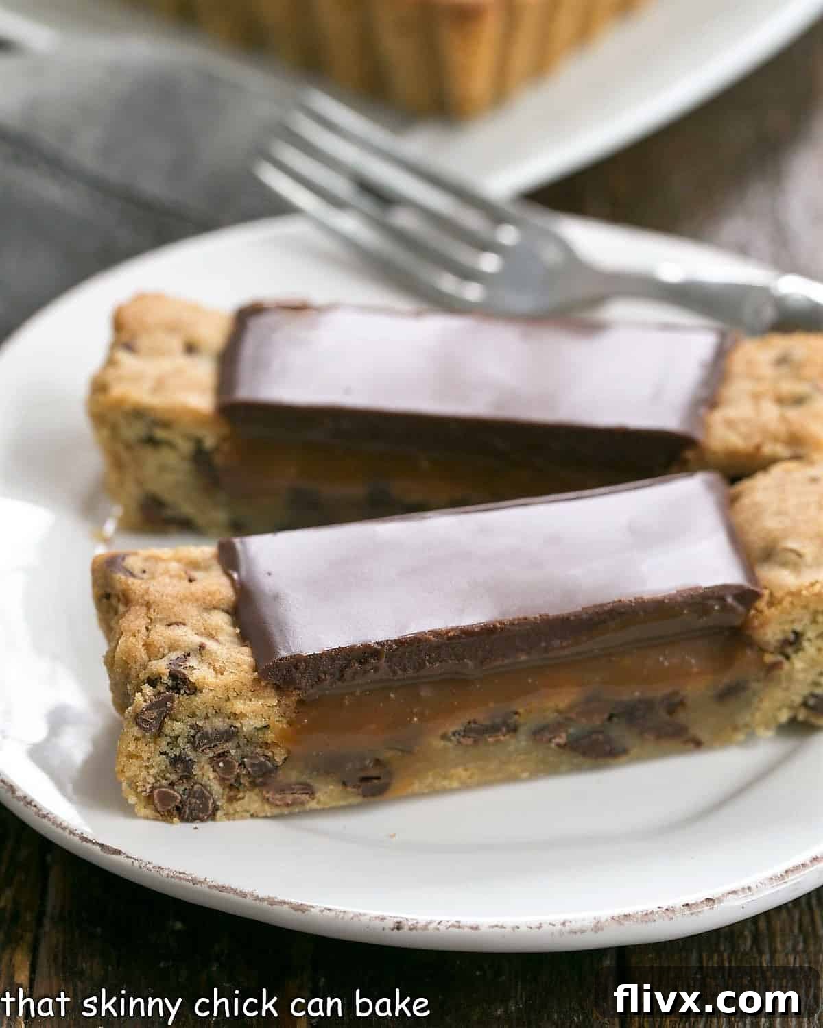 Two slices of chocolate chip cookie tart on a white plate with a fork, showing layers of cookie, caramel, and chocolate glaze.