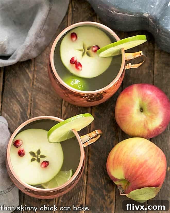 An inviting overhead view of two perfectly crafted Apple Cider Moscow Mules in elegant copper mugs, adorned with vibrant apple slices, lime wedges, and scattered pomegranate seeds, set against a warm, autumnal backdrop.