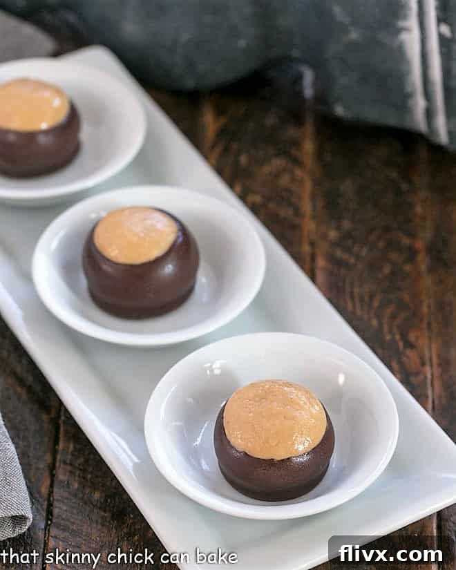 Three perfectly crafted Chocolate Peanut Butter Bon Bons, partially dipped to reveal their peanut butter core, are artfully arranged in individual white bowls on a sleek white serving tray, showcasing their elegant simplicity and irresistible appeal.