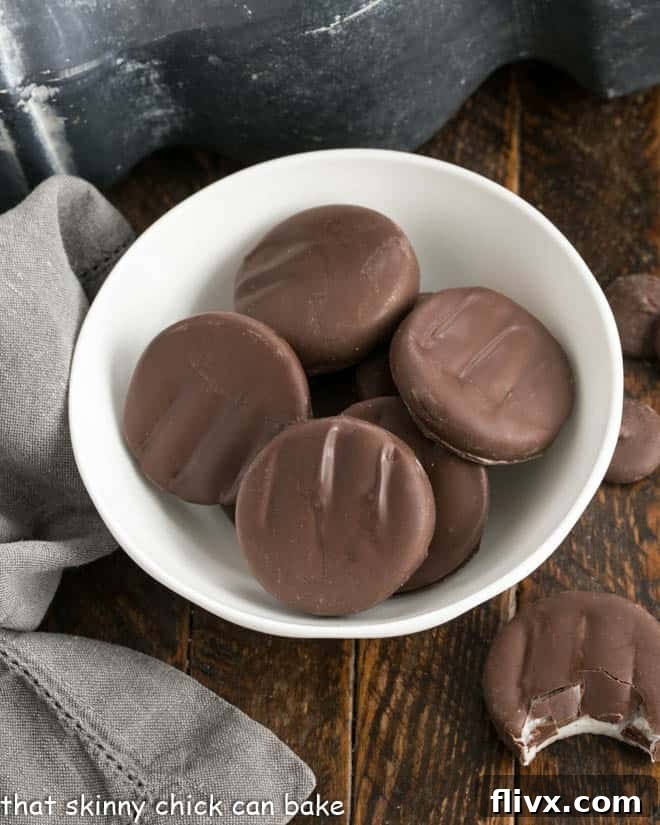 Overhead view of several homemade Peppermint Patties in a white bowl, with one patty on the counter having a bite removed, revealing the white minty filling.