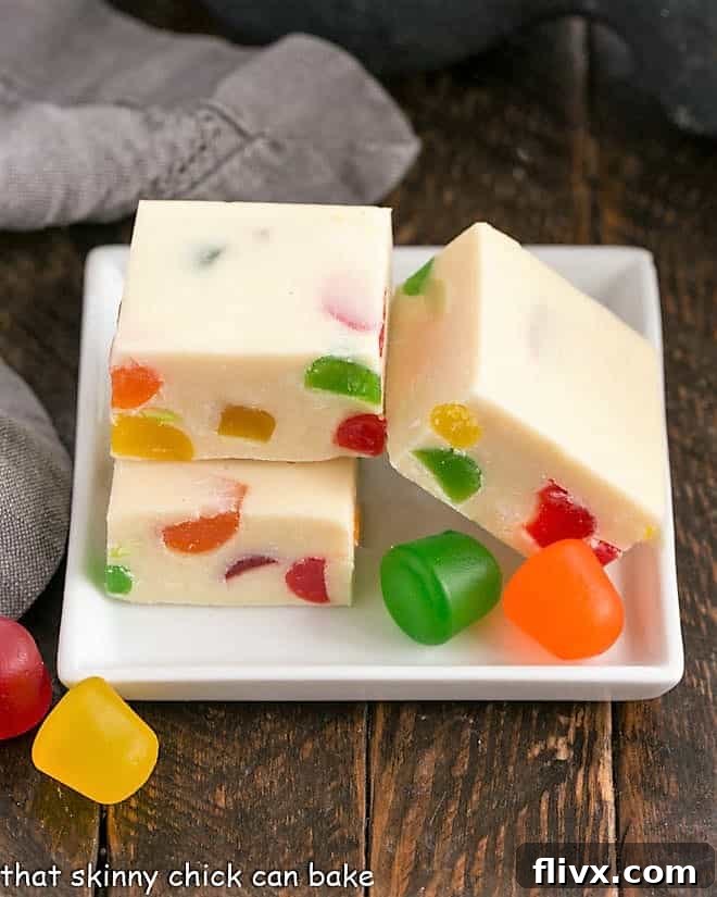 Three cubes of festive white chocolate fudge on a square white plate, garnished with colorful gumdrops, ready for a holiday treat.