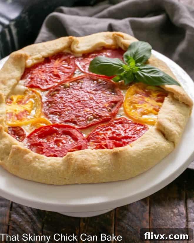 A beautifully baked Cheese and Tomato Galette resting on a white cake stand, garnished with fresh basil leaves. The golden crust and vibrant red tomato slices are clearly visible, promising a delicious savory treat.