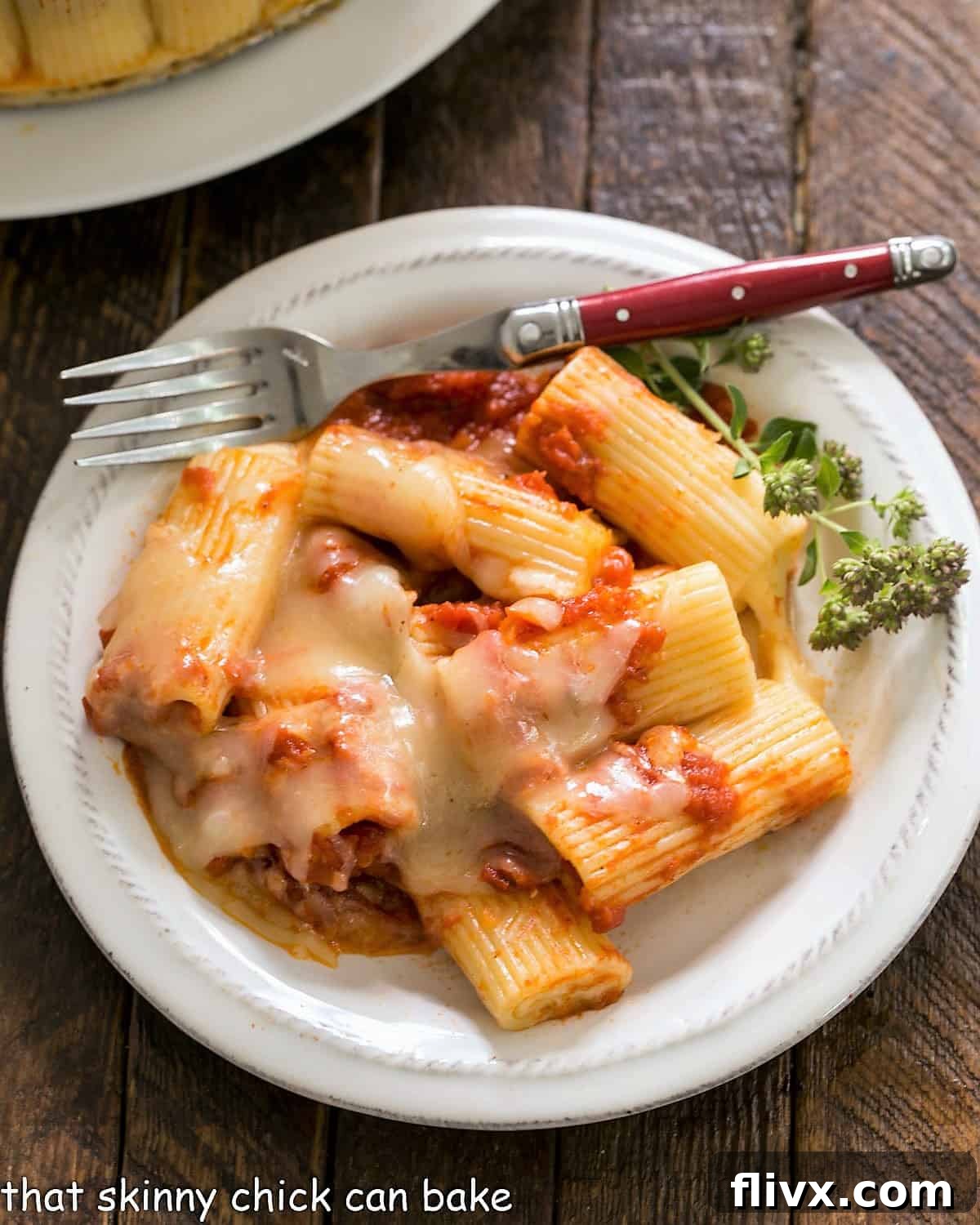 Overhead view of baked rigatoni on a small white plate with a fork and herb garnish.