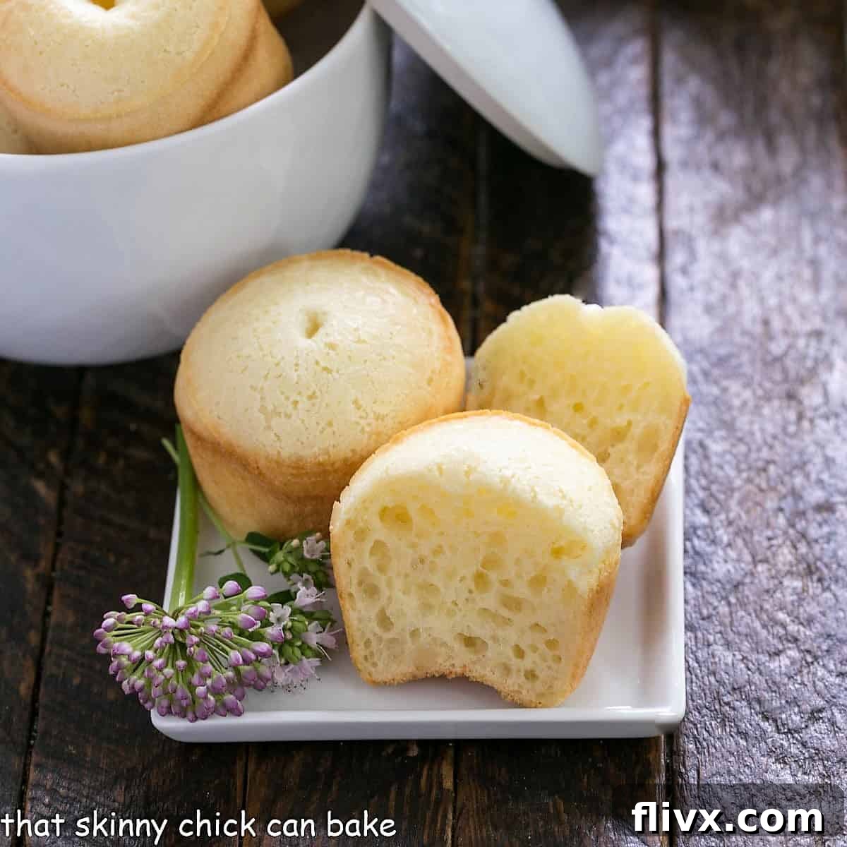 A selection of freshly baked Brazilian cheese bread, some sliced open to reveal the soft, airy interior, garnished with a chive flower on a white plate.