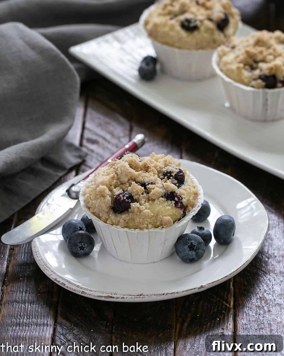 A single blueberry cream cheese muffin on a white plate, cut open to reveal the creamy filling, accompanied by a knife and fresh blueberries.