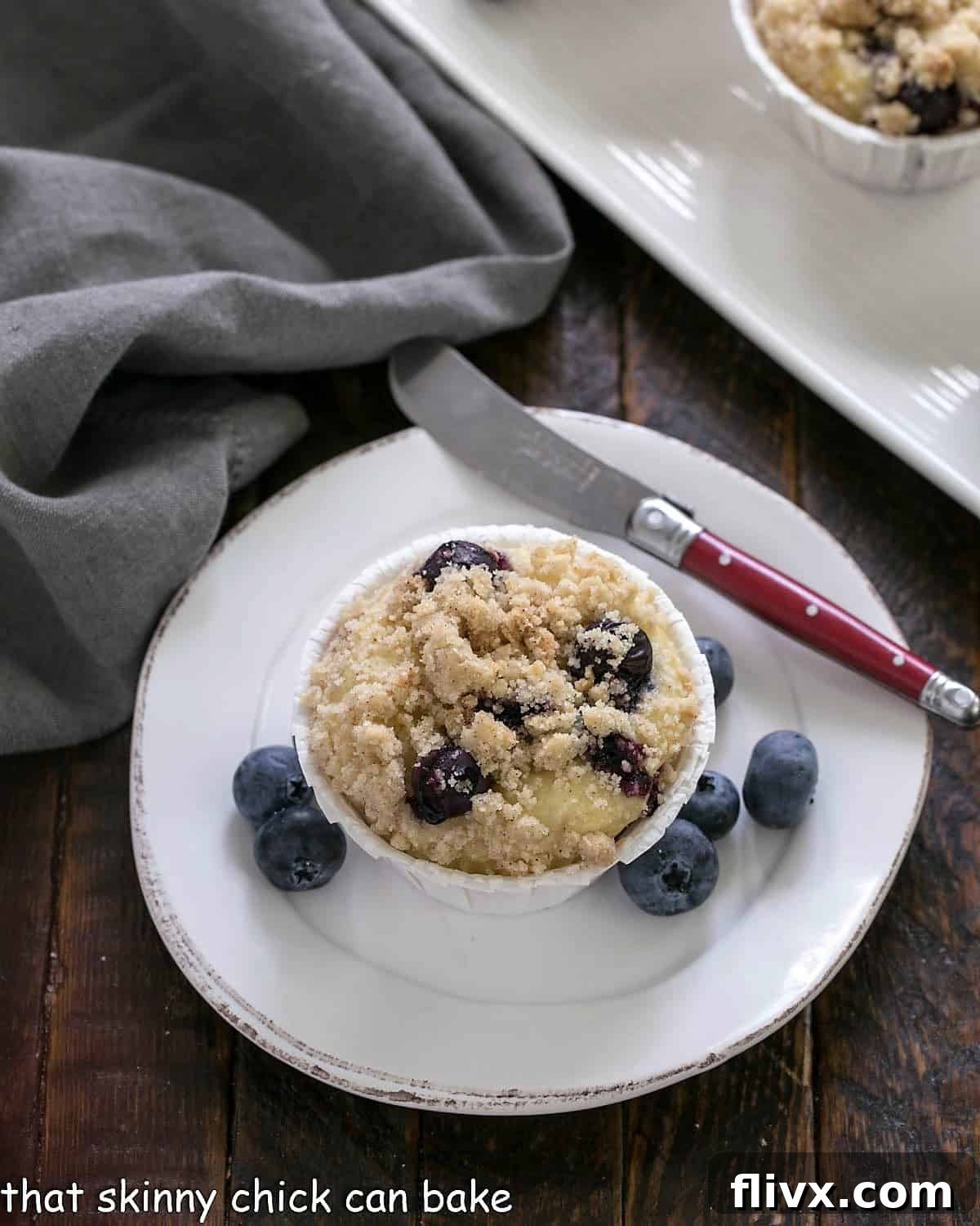 An overhead view of a single blueberry muffin on a white plate, with a vintage-style red handle knife.