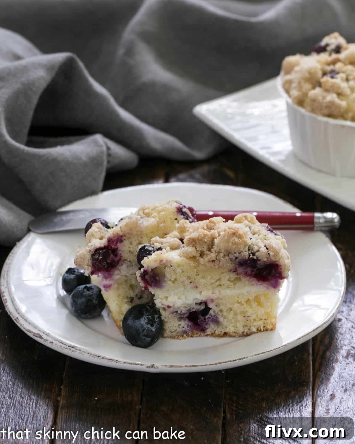 Two halves of a blueberry streusel muffin, revealing the moist interior and layers of blueberries and cream cheese filling, on a white round plate.