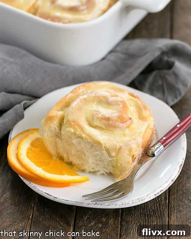 Homemade Orange Yeast Rolls - a delicious twist on cinnamon rolls filled and iced with orange frosting! Homemade Orange Sweet Rolls nestled in a baking dish, with one roll elegantly placed on a small plate beside a red-handled fork, ready to be enjoyed.