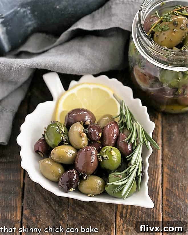 Close-up of herbed olives in a ceramic leaf plate, garnished with a slice of lemon and fresh rosemary.