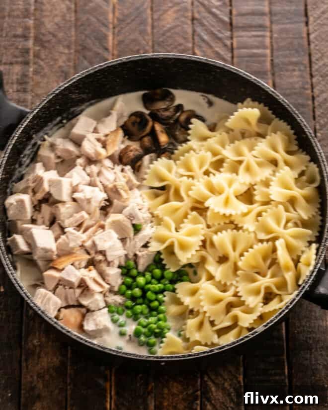 Cooked pasta, tender turkey, sautéed mushrooms, and vibrant peas are carefully folded into the creamy sauce within a large skillet, ready for baking.