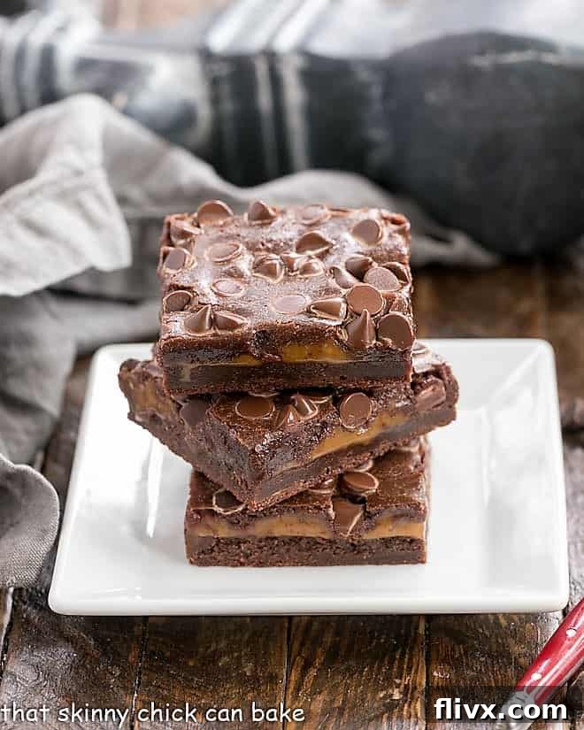 A close-up view of a layered caramel brownie resting on a square white plate, showcasing the rich, dark brownie layers sandwiching a thick, glossy caramel center.