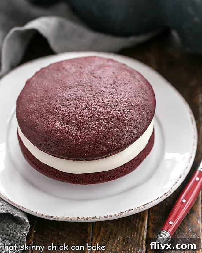 A single, perfectly crafted red velvet whoopie pie sits invitingly on a small, round white plate, highlighting its luscious filling.