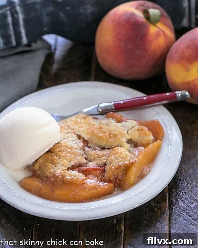 A serving of homemade peach cobbler on a white plate, topped with melting vanilla ice cream, with two fresh, unpeeled peaches in the soft-focused background.