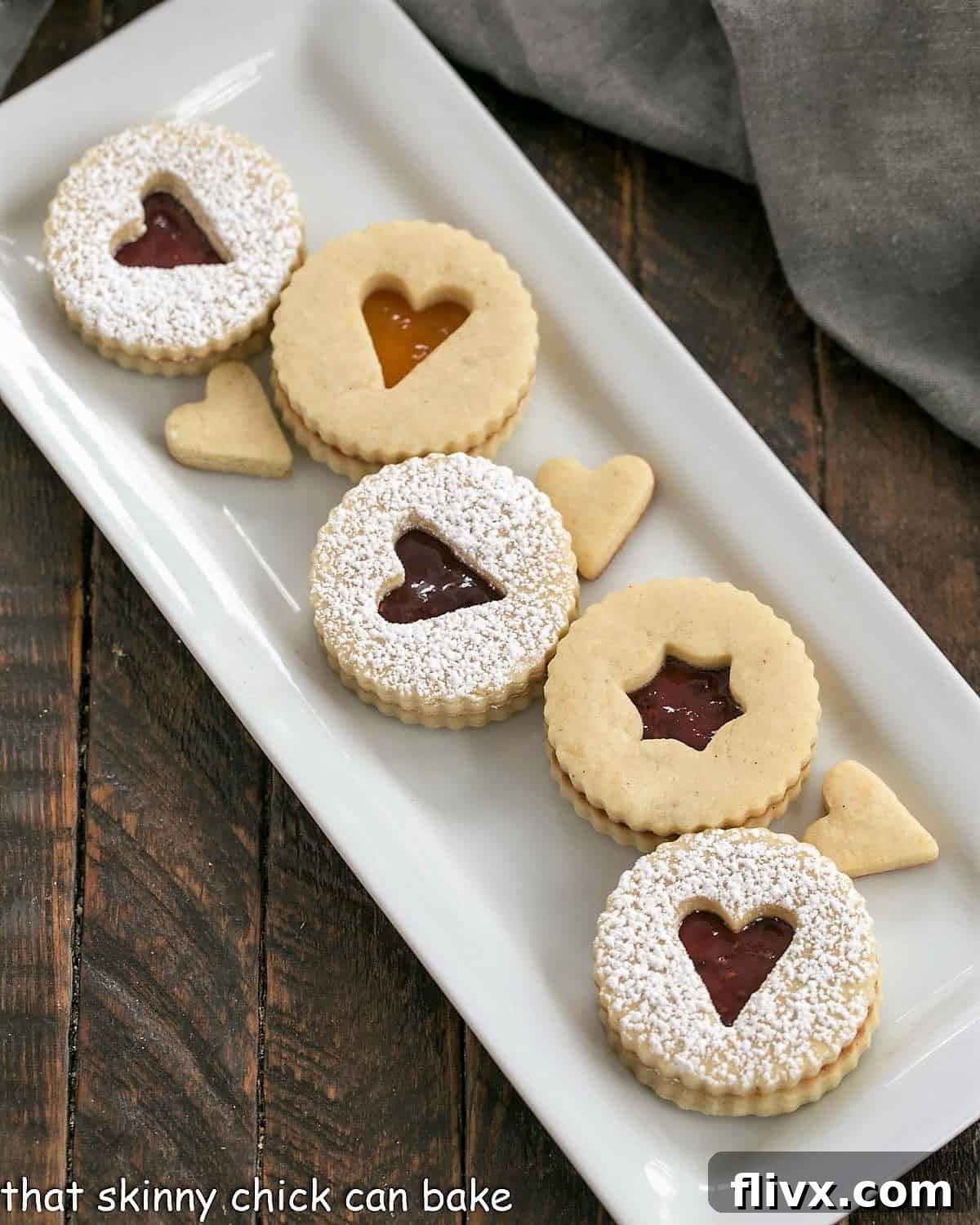 Overhead view of linzer cookies on a white ceramic tray, showcasing their delicate powdered sugar dusting and visible jam filling through the cutout.