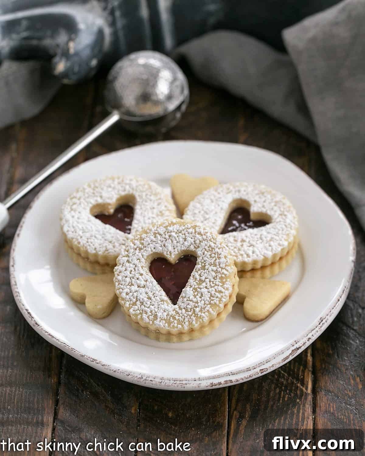 3 linzer cookies dusted with powdered sugar on a round white dessert plate, highlighting the raspberry jam filling.